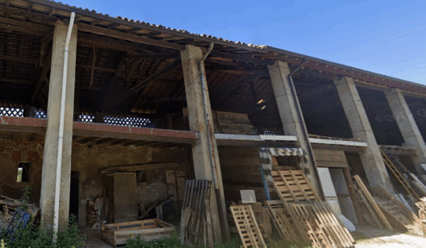 Struttura di cascina con pilastri, tetto in legno e materiali accatastati sul lato.
