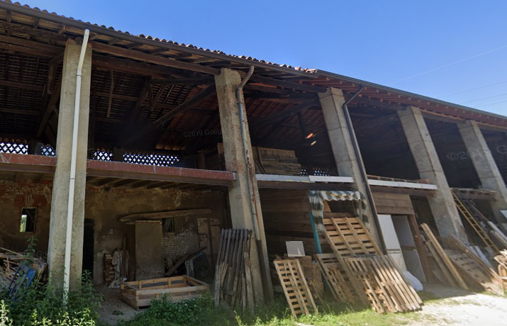Struttura di cascina con pilastri, tetto in legno e materiali accatastati sul lato.